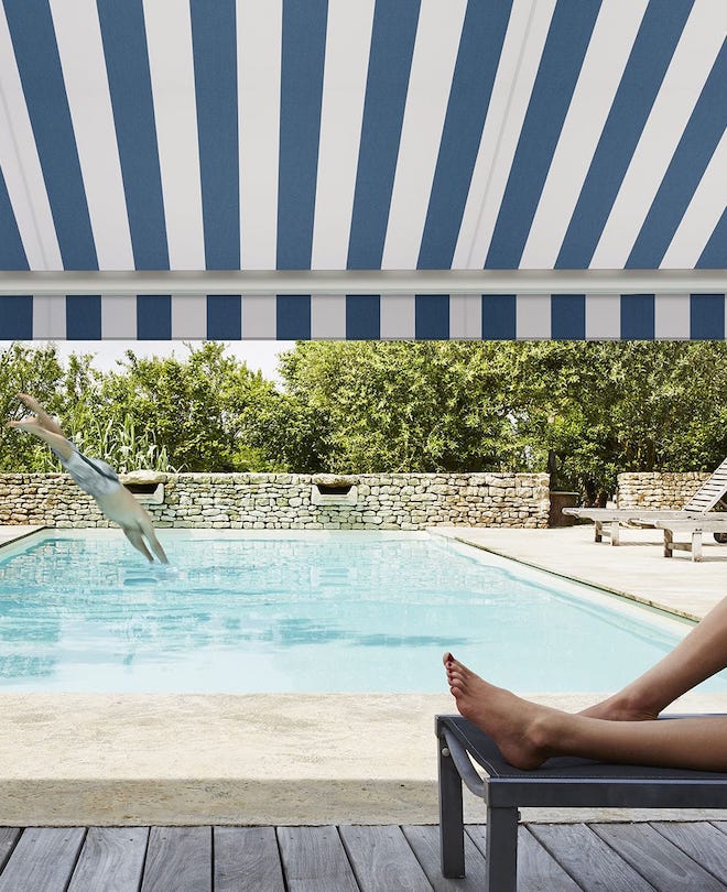 A blue striped awning with a diver diving into a pool beyond and a woman reclined beneath