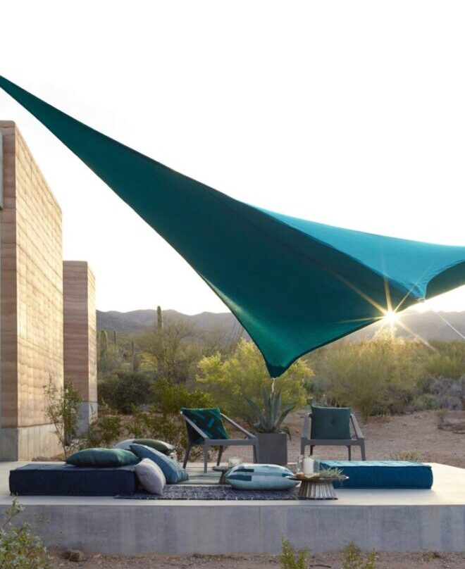 a blue sun shade over a patio with two chairs, a plant, a rug, and several cushions