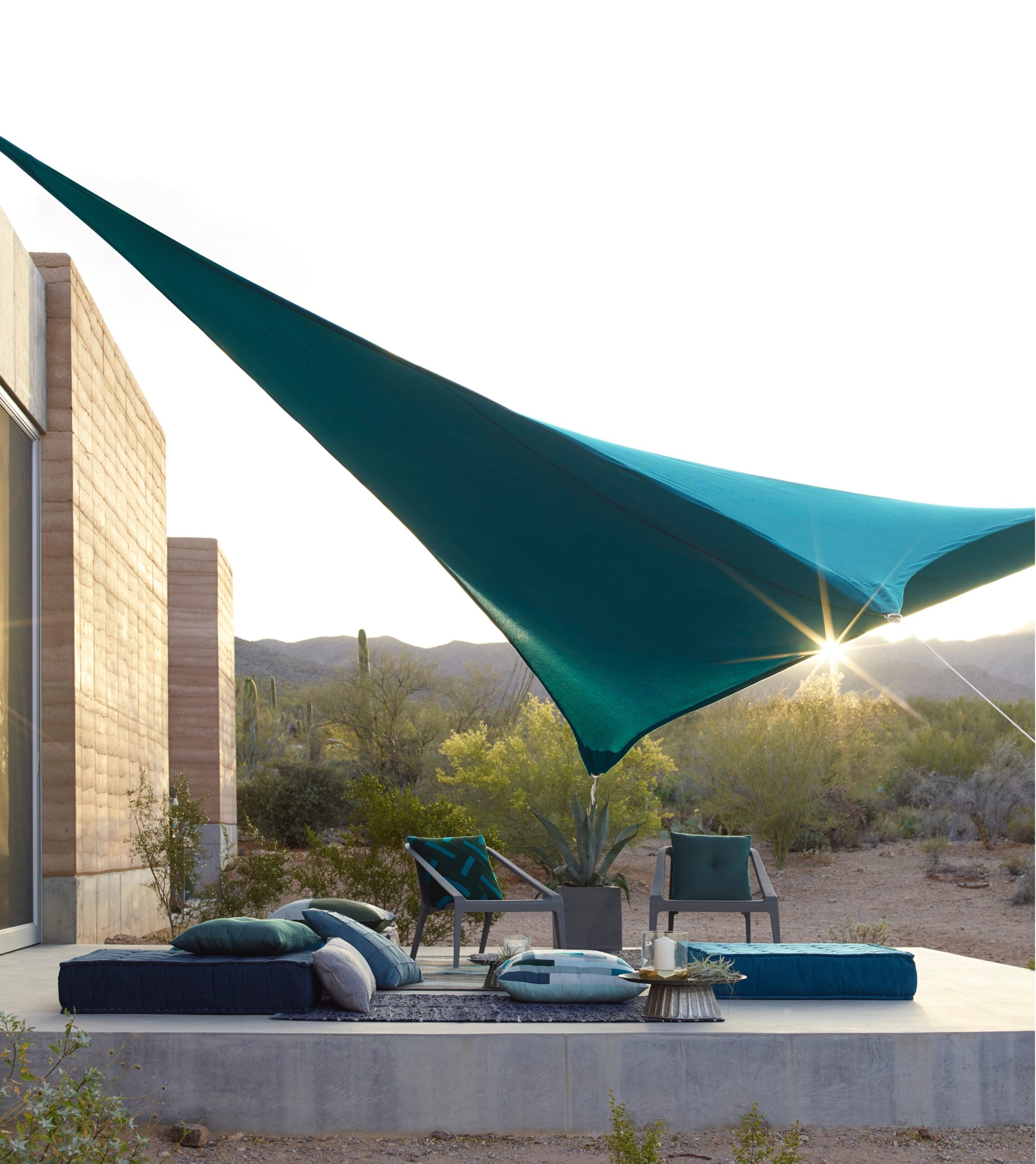 a blue sun shade over a patio with two chairs, a plant, a rug, and several cushions