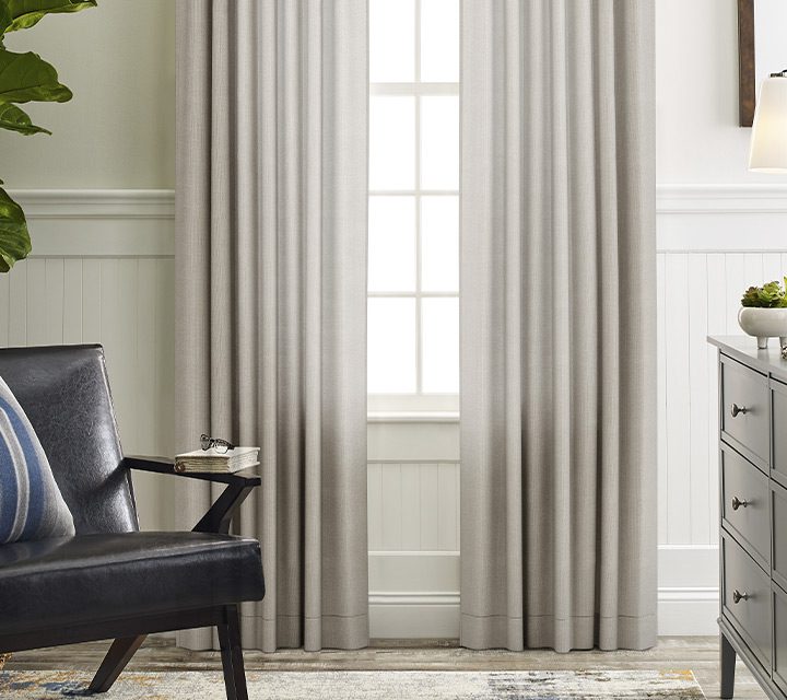 A room with neutral window coverings flanked by a chair, plant, and other furniture