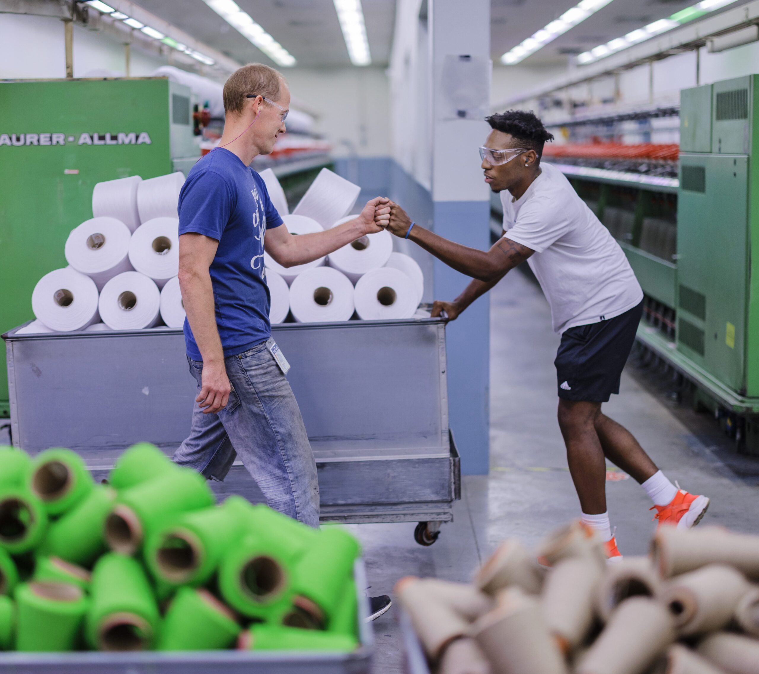 two men fist bump in front of large containers full of thread