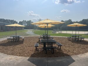 Black picnic tables sit beneath yellow umbrellas on a bed of mulch beside a baseball field