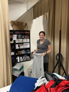 Dark haired woman in a striped v neck tshirt stands indoors next to a shelf, folds a gray tshirt, and smiles at the camera